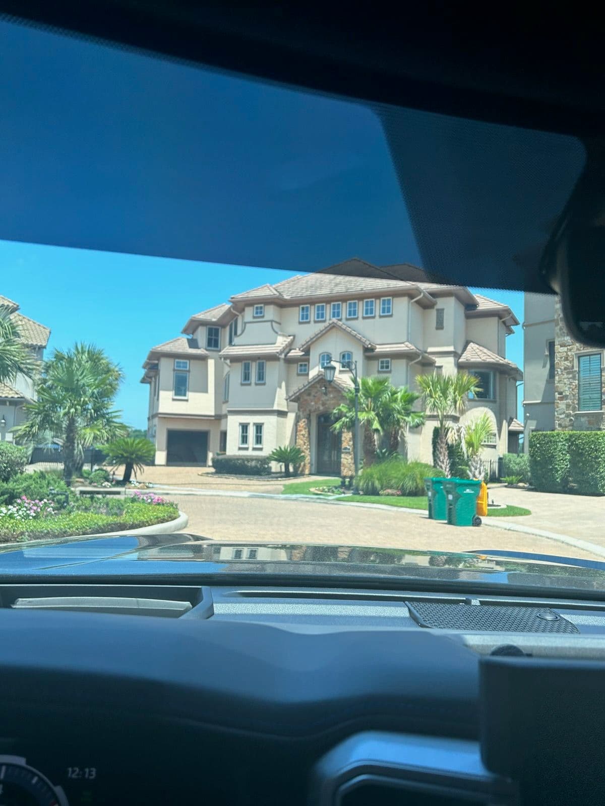 Luxurious mansion with palm trees and manicured landscaping viewed from a car dashboard.